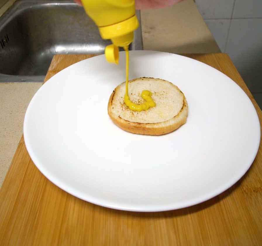A hand squeezing bright yellow mustard from a plastic squeeze bottle onto a toasted burger bun resting on a white plate.