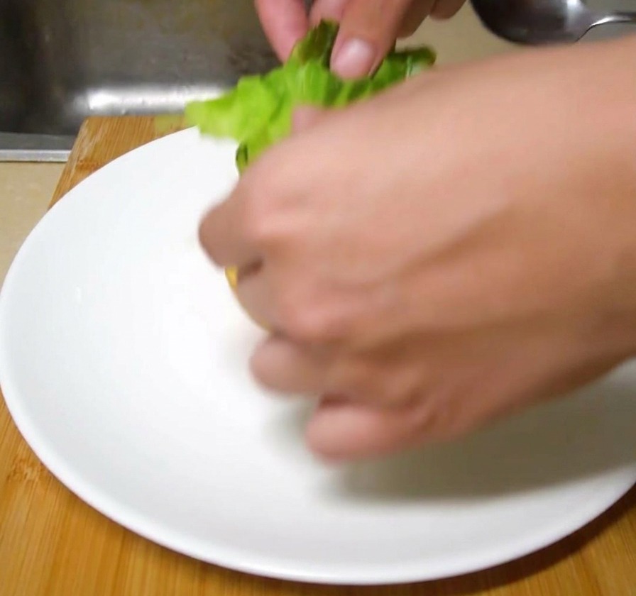 Fresh green lettuce leaves being hand-placed onto a toasted burger bun on a white ceramic plate.