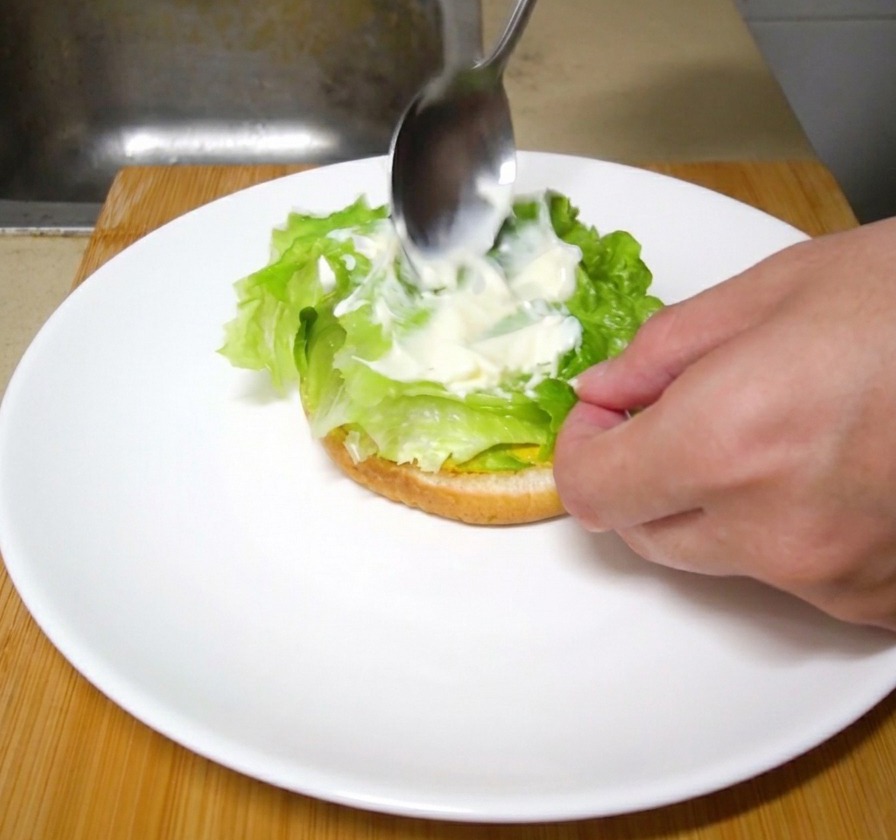 A metal spoon spreading a dollop of white mayonnaise over fresh green lettuce leaves on a burger stack.