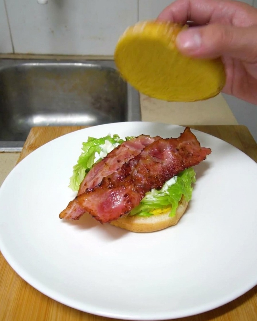 A hand lowering the top burger bun onto a plated burger consisting of lettuce and crispy bacon strips.