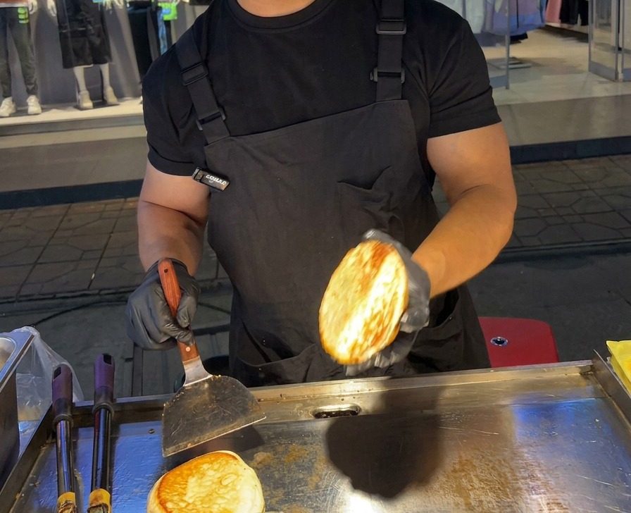 A person holding up a perfectly toasted, golden-brown burger bun half over a flat stainless steel griddle.