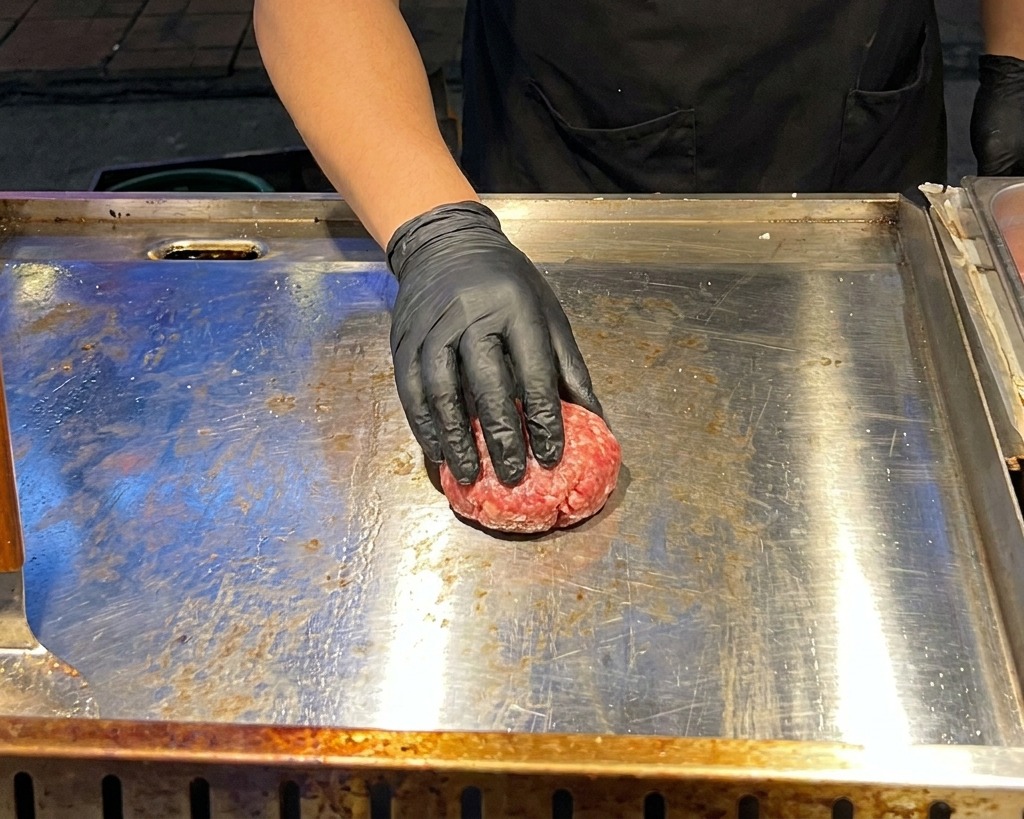 A hand in a black glove using a flat metal press to squash a raw beef meatball into a patty shape on a hot griddle.