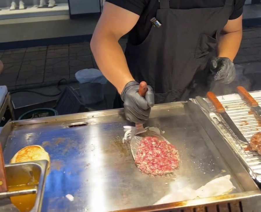 A metal spatula flipping a beef patty over a pile of grilled purple onions on a commercial griddle.