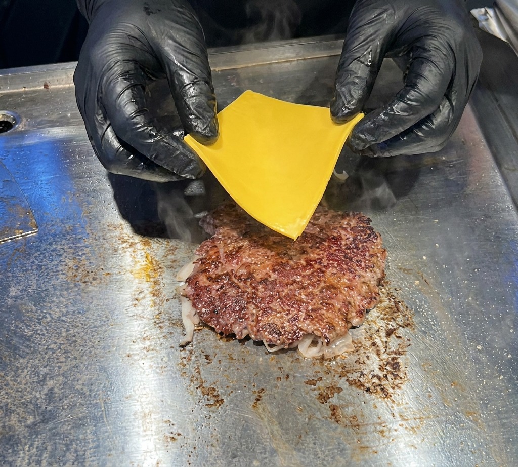 A close-up of black-gloved hands carefully placing a slice of bright yellow American cheese onto a sizzling, browned beef patty on a flat griddle.