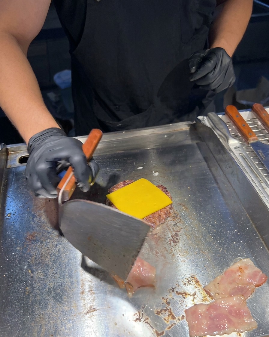 A metal spatula flipping sizzling bacon strips on a griddle next to a beef patty topped with melted yellow cheese.