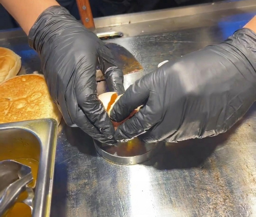 Black-gloved hands cracking a fresh egg into a silver circular metal mold resting on a hot stainless steel cooking surface.