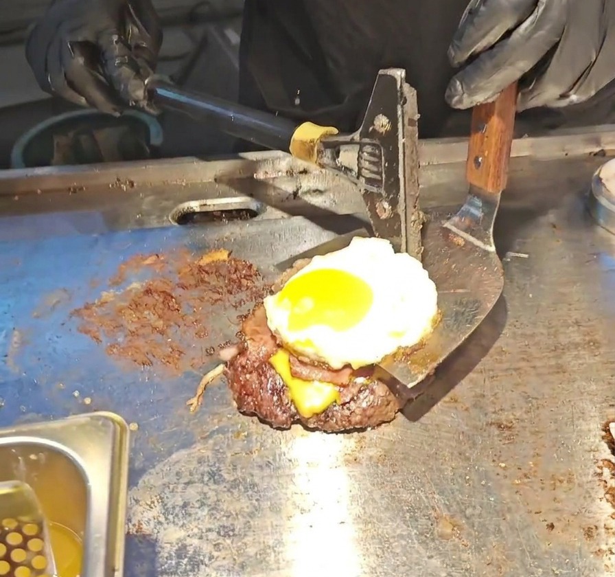 A wide metal spatula lifting a tall burger stack consisting of a beef patty, melted cheese, bacon, and a runny sunny-side-up egg.