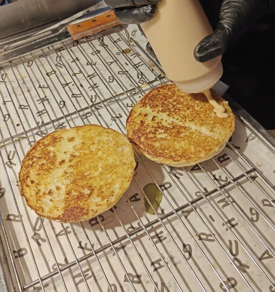 Creamy orange-colored sauce being squeezed from a plastic bottle onto two toasted bun halves resting on a wire rack.