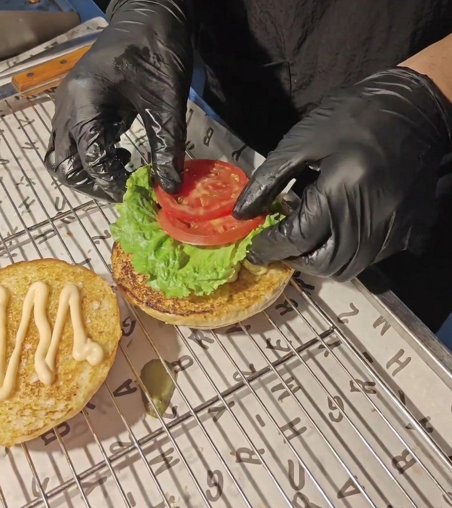Hands in black gloves placing fresh tomato slices onto a green lettuce leaf on a toasted burger bun.