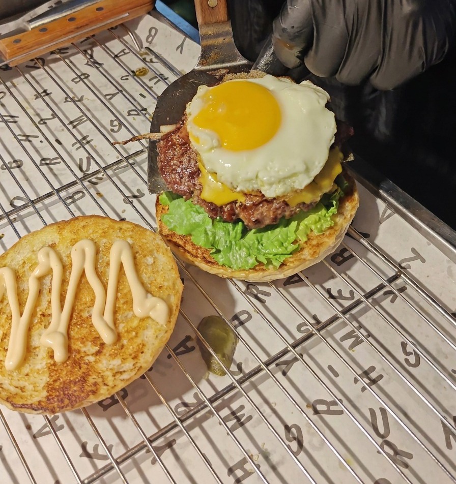 A metal spatula transferring a beef patty topped with melted cheese and a sunny-side-up egg onto the lettuce and tomato layer.