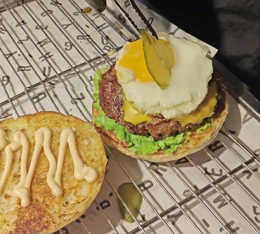 Tongs placing a thin slice of pickled cucumber onto a bright yellow egg yolk on top of a burger stack.