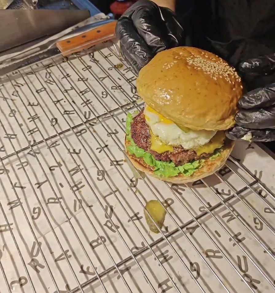 Hands in black gloves placing a golden sesame seed bun on top of a fully assembled bacon and egg burger.