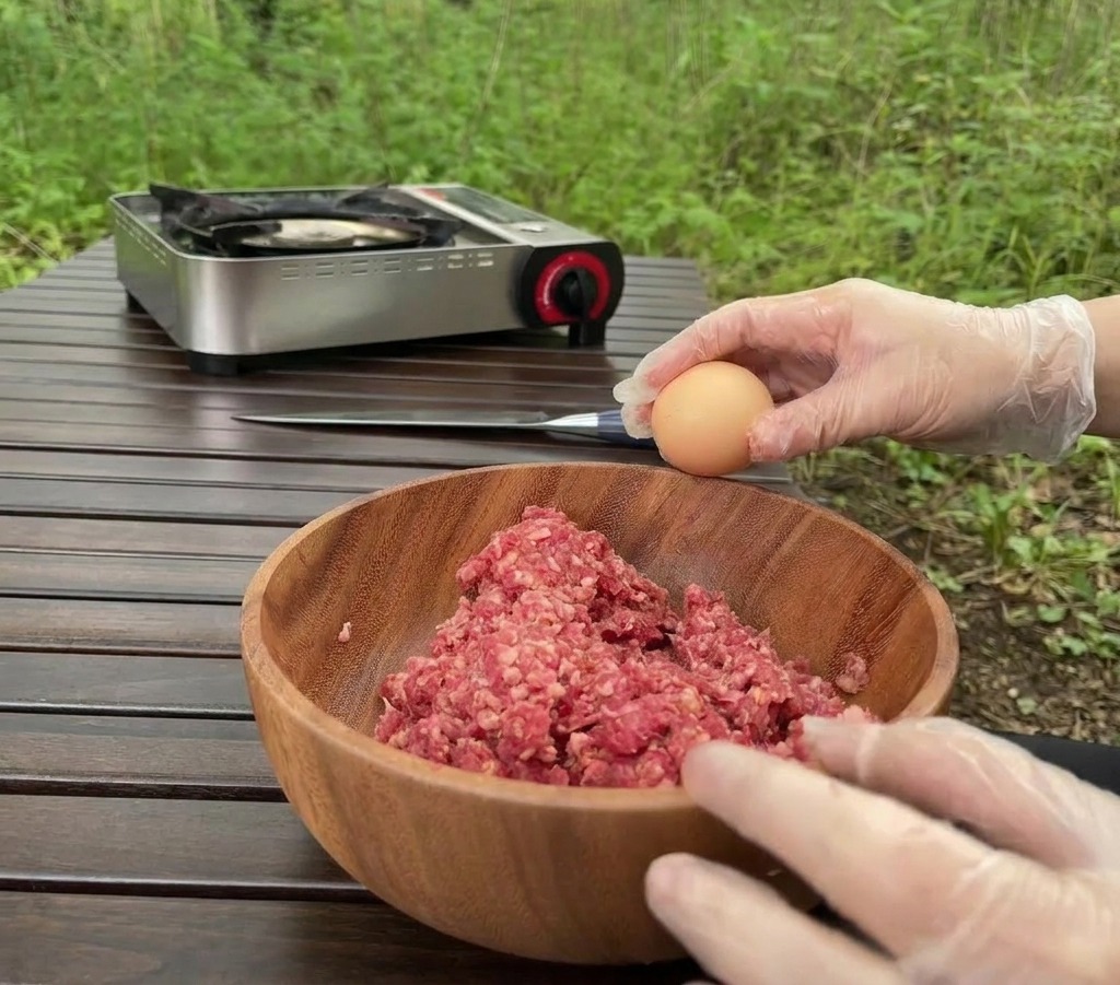 A person wearing clear gloves holding a brown egg over a wooden bowl filled with raw ground beef on a wooden table outdoors.