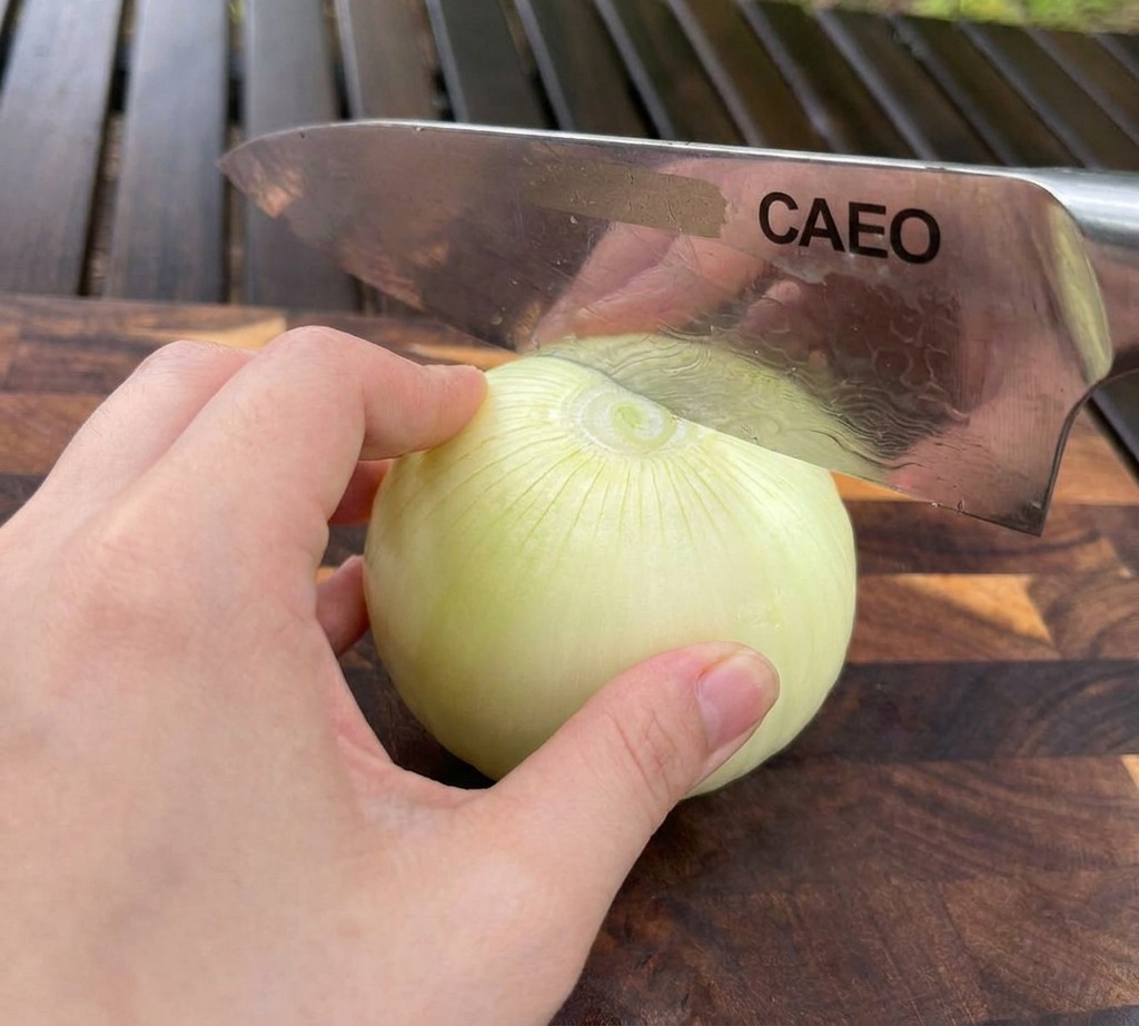A hand stabilizing a white onion on a wooden cutting board while a chefs knife slices through the top.