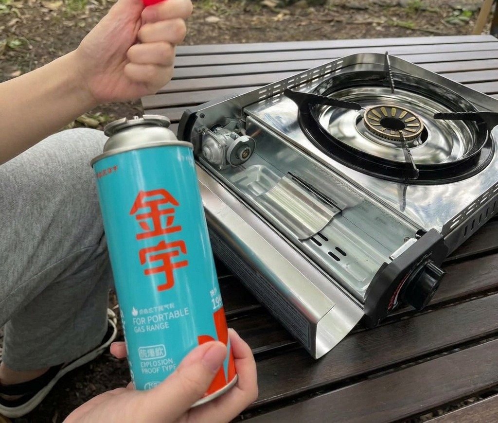 A person holding a blue butane gas canister and its red cap next to an open portable stove compartment.