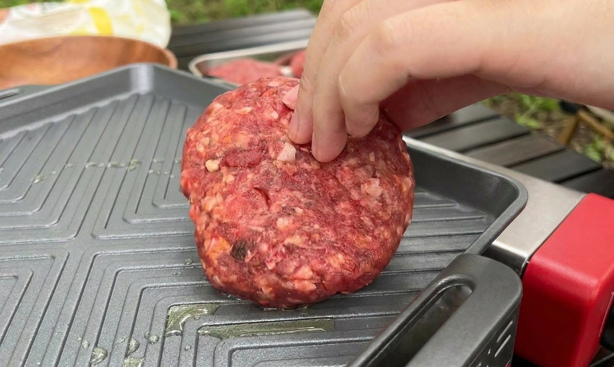 A hand placing a thick, raw beef patty onto a hot, ridged square griddle pan during an outdoor cooking session.