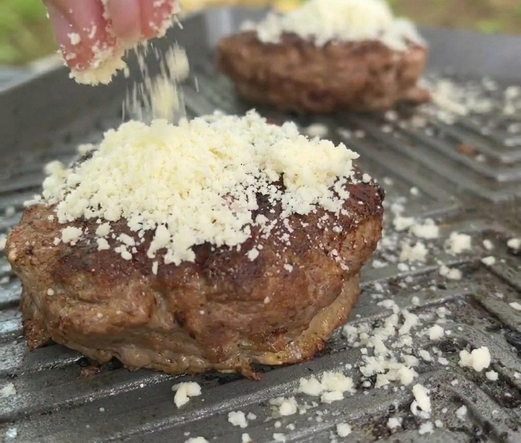 A close-up of shredded white cheese being sprinkled onto a browned beef patty on a hot griddle pan.