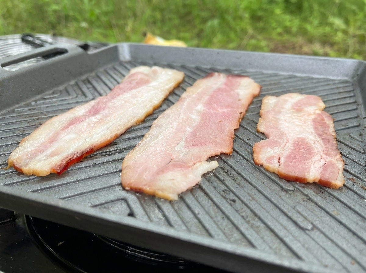Three long strips of bacon frying on a grey ridged griddle pan, showing the fat rendering.