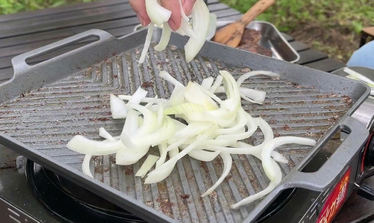 Thinly sliced white onions being dropped onto a hot griddle pan to be sautéed next to the meat.
