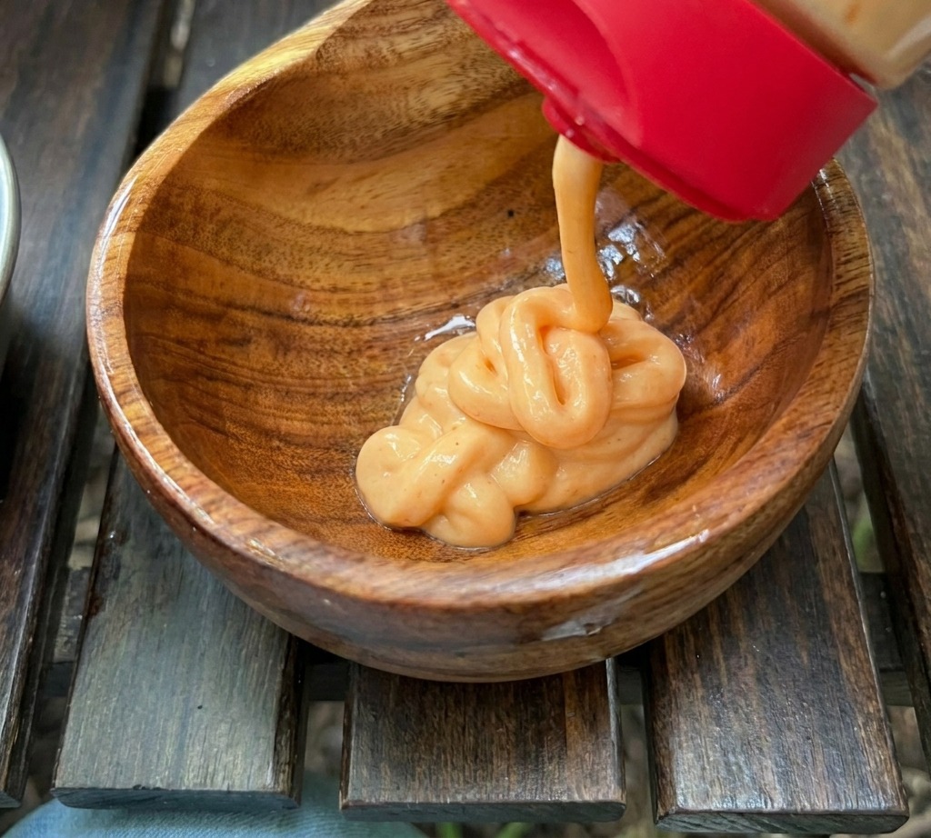 Creamy orange sauce being squeezed from a red-capped bottle into a small wooden bowl containing other sauce ingredients.