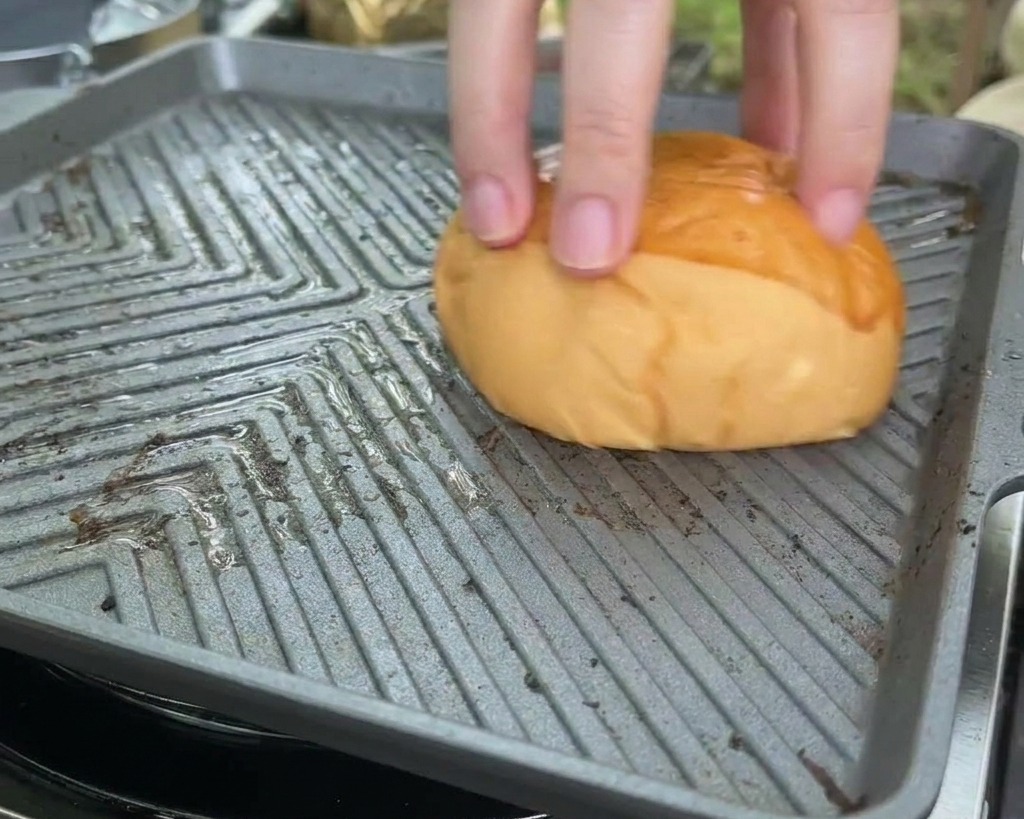 A brioche bun being pressed face-down onto a hot, square griddle pan to toast the cut side.