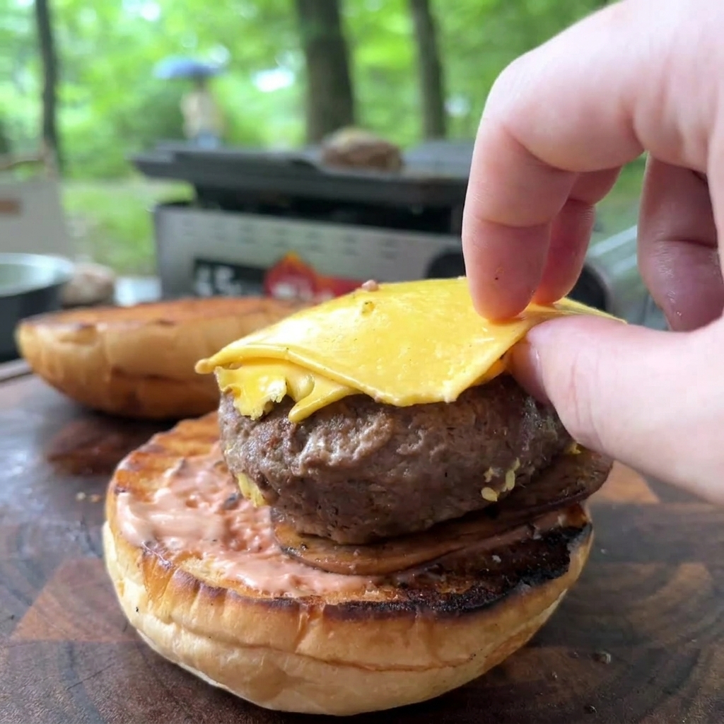 A hand placing a cooked beef patty with melted yellow cheese onto a toasted bun spread with pink sauce.