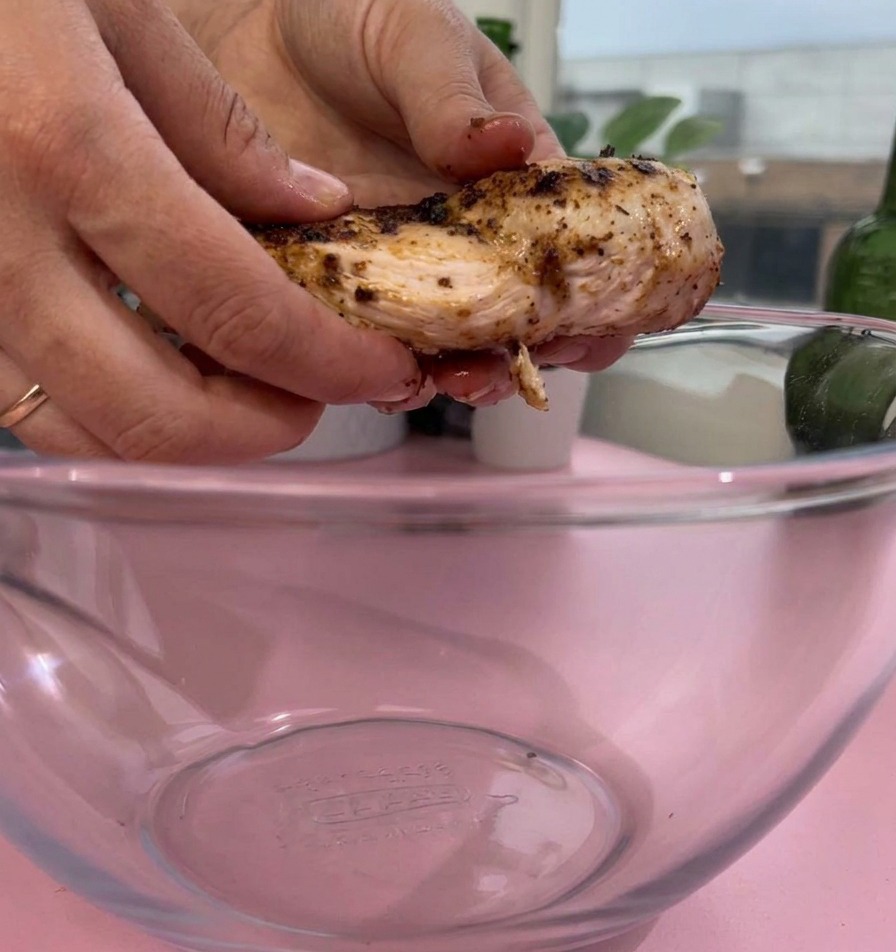 A pair of hands placing a piece of cooked, seasoned chicken breast into a clear glass mixing bowl on a pink surface.