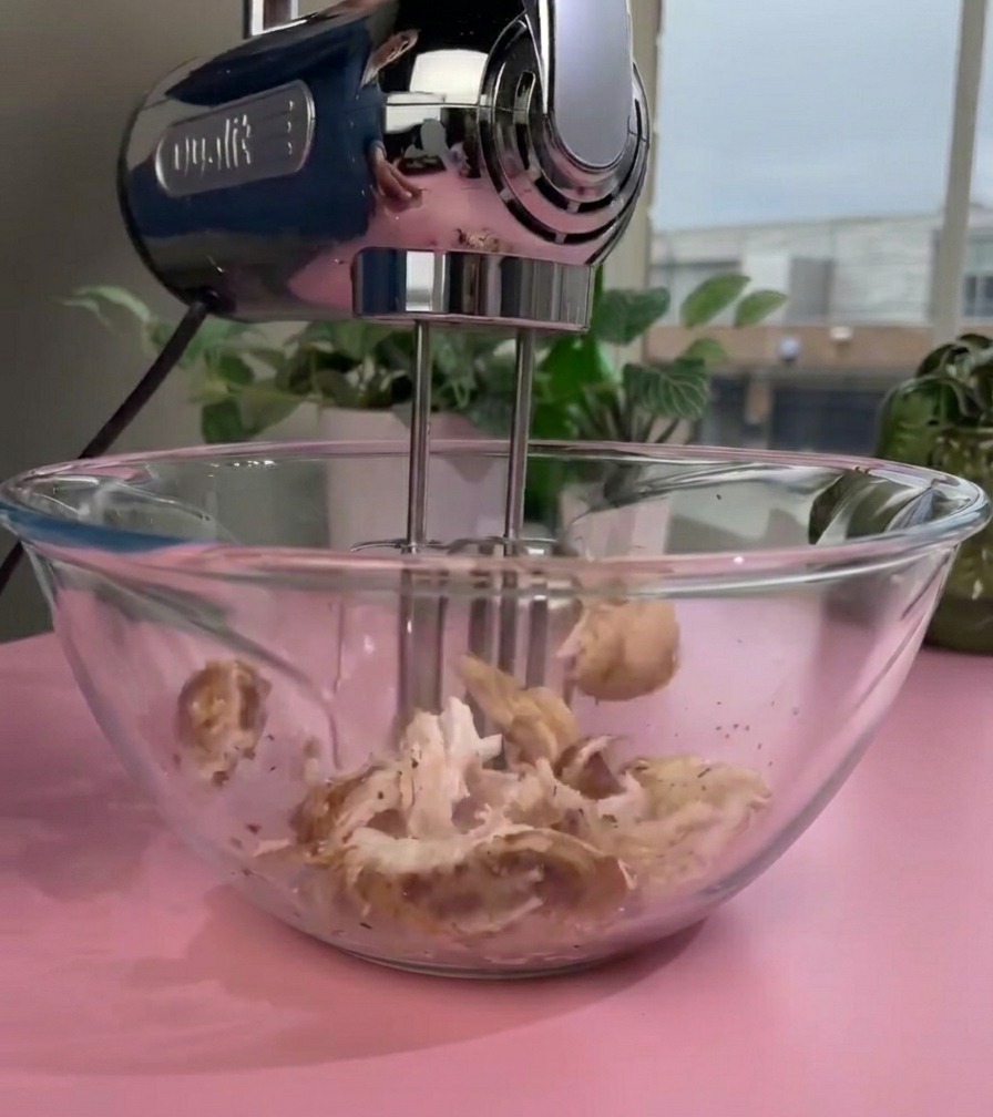 An electric hand mixer with rotating beaters shredding cooked chicken inside a glass bowl creating a fine texture