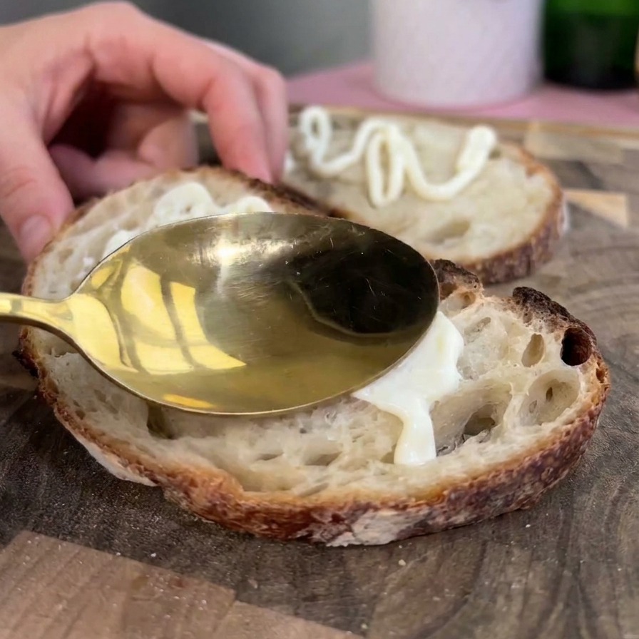 A gold spoon spreading thick white mayonnaise onto a toasted slice of rustic sourdough bread.