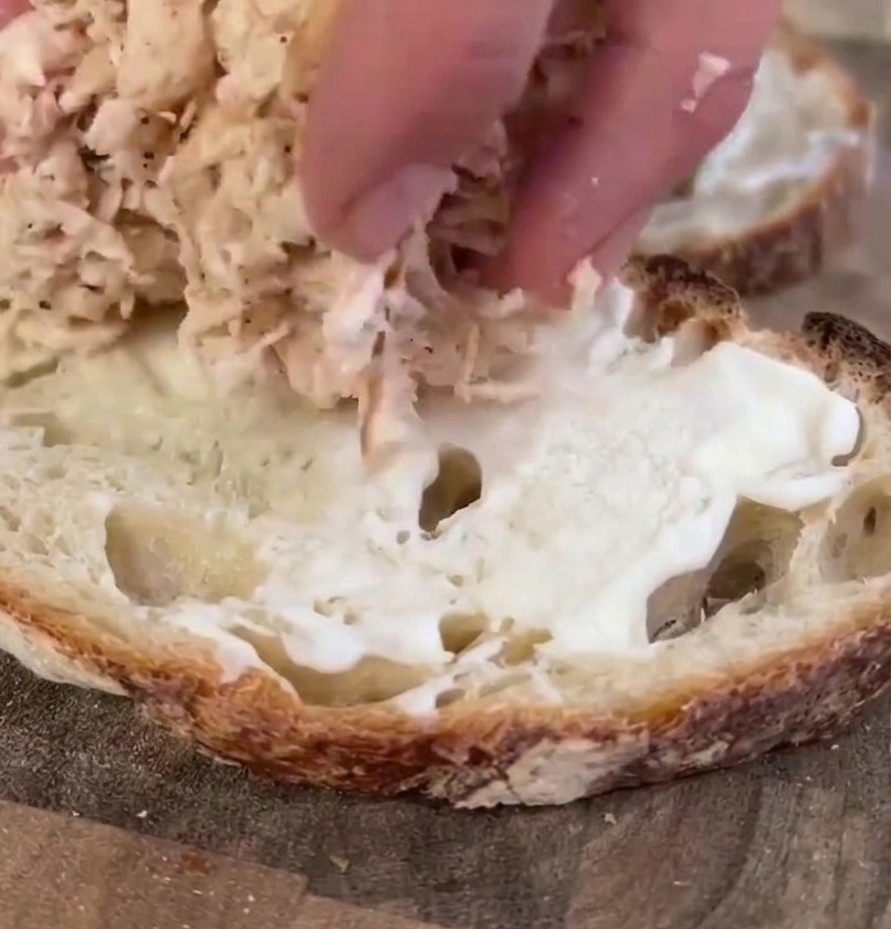 A persons hand placing a mound of shredded cooked chicken onto a slice of sourdough bread prepared with mayonnaise.