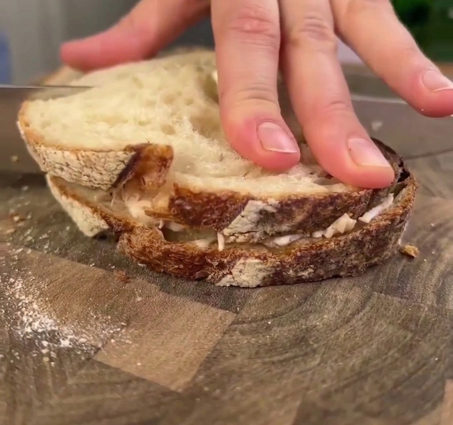 A hand pressing down firmly on a large sourdough chicken sandwich on a wooden board while a knife prepares to cut it.