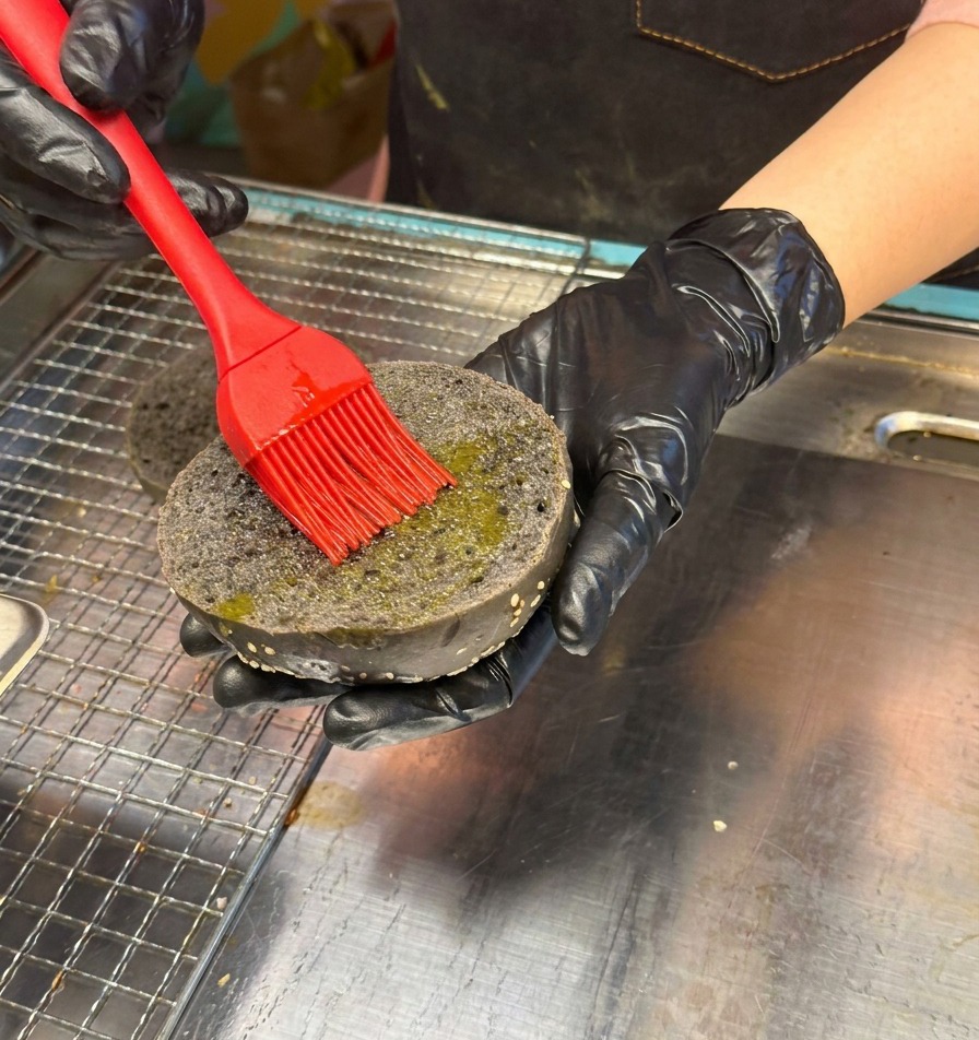A hand in a black glove using a red silicone brush to apply oil to the inner face of a black sesame burger bun.