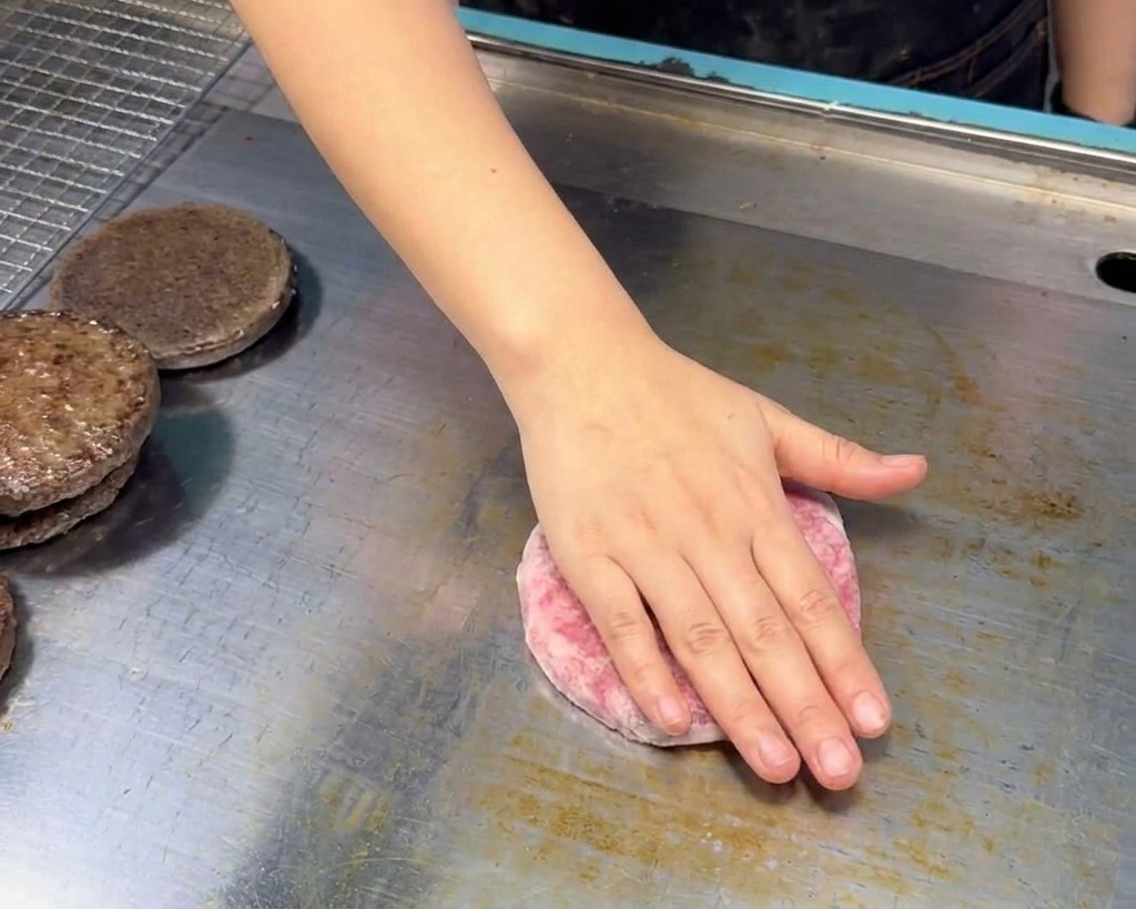 A hand placing a raw, pink grain-fed beef patty onto a clean stainless steel griddle surface next to toasted bun halves.