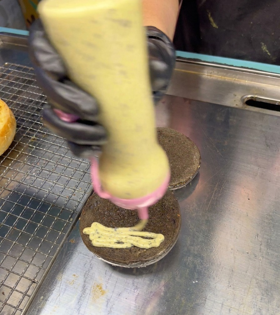 A squeeze bottle applying a creamy, speckled truffle sauce in a zig-zag motion onto a black burger bun.