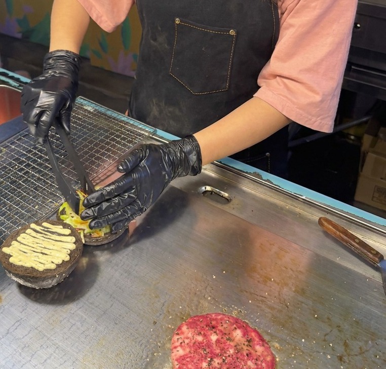 Tongs dropping a mixture of shredded lettuce and purple cabbage onto a black bun already coated with truffle sauce