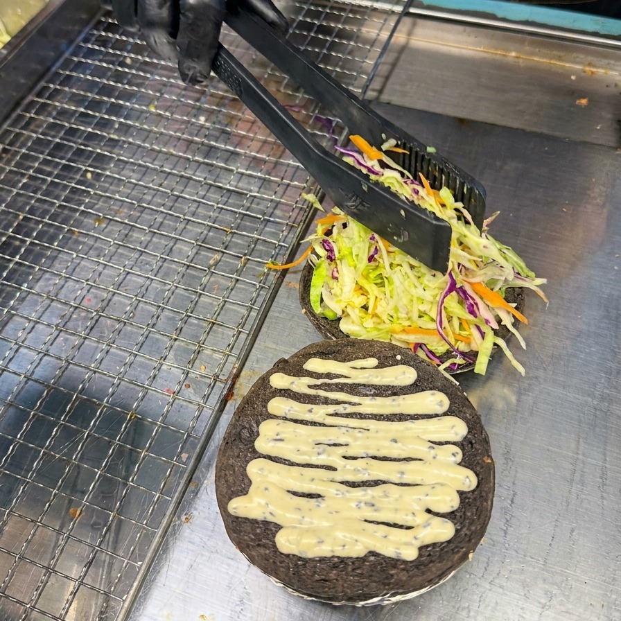 A person using tongs to carefully place a bright red tomato slice onto a bed of greens on a charcoal-black burger bun