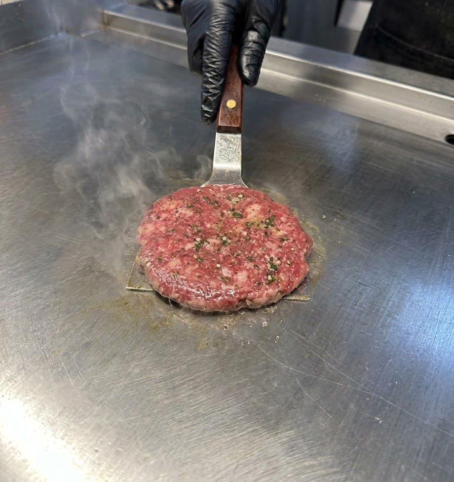 A metal spatula pressing down on a beef patty on a stainless steel griddle with steam rising.