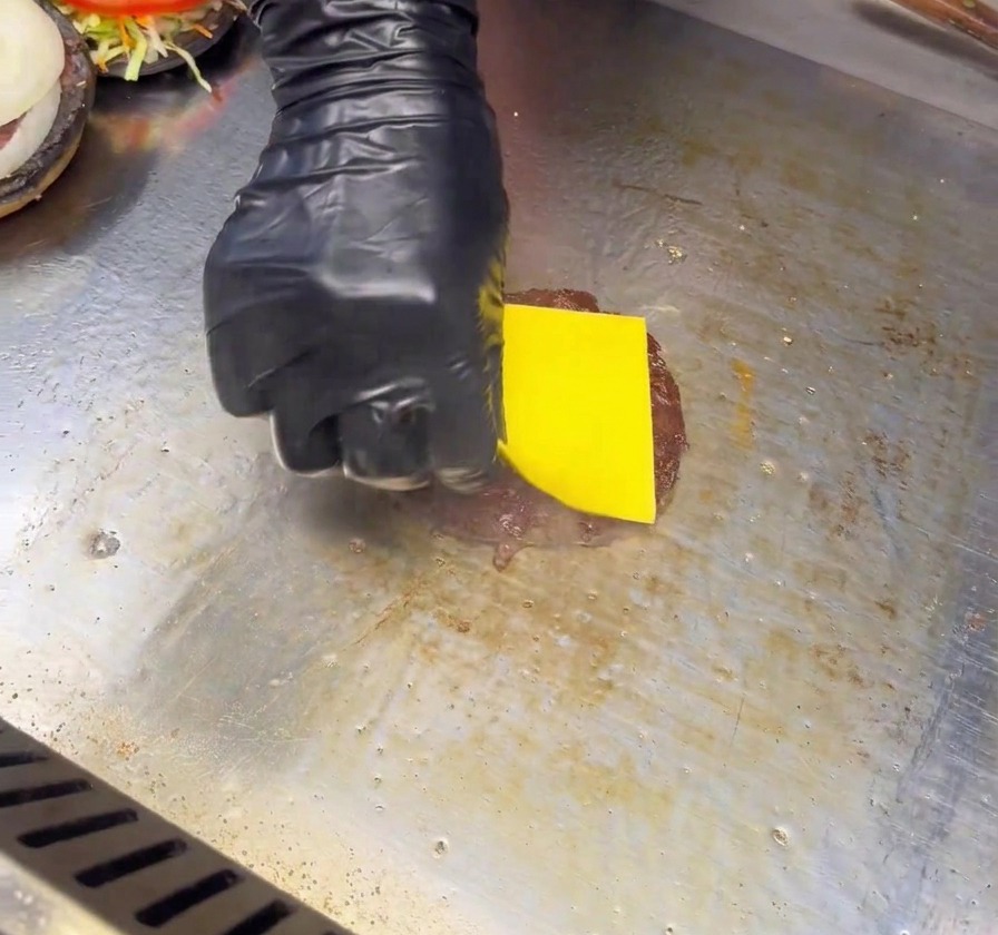 A hand in a black glove placing a yellow cheese slice onto a seared burger patty on a flat-top grill.