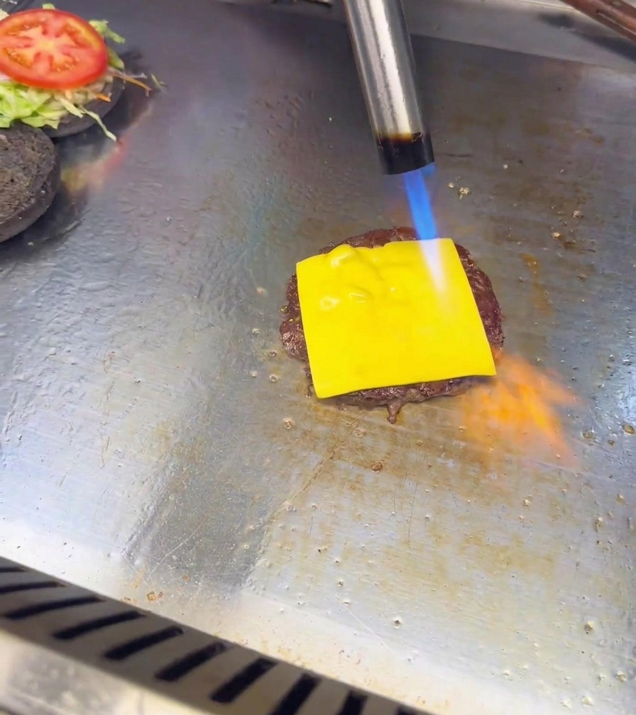 A blue and orange flame from a kitchen torch melting a slice of cheese on a burger patty.