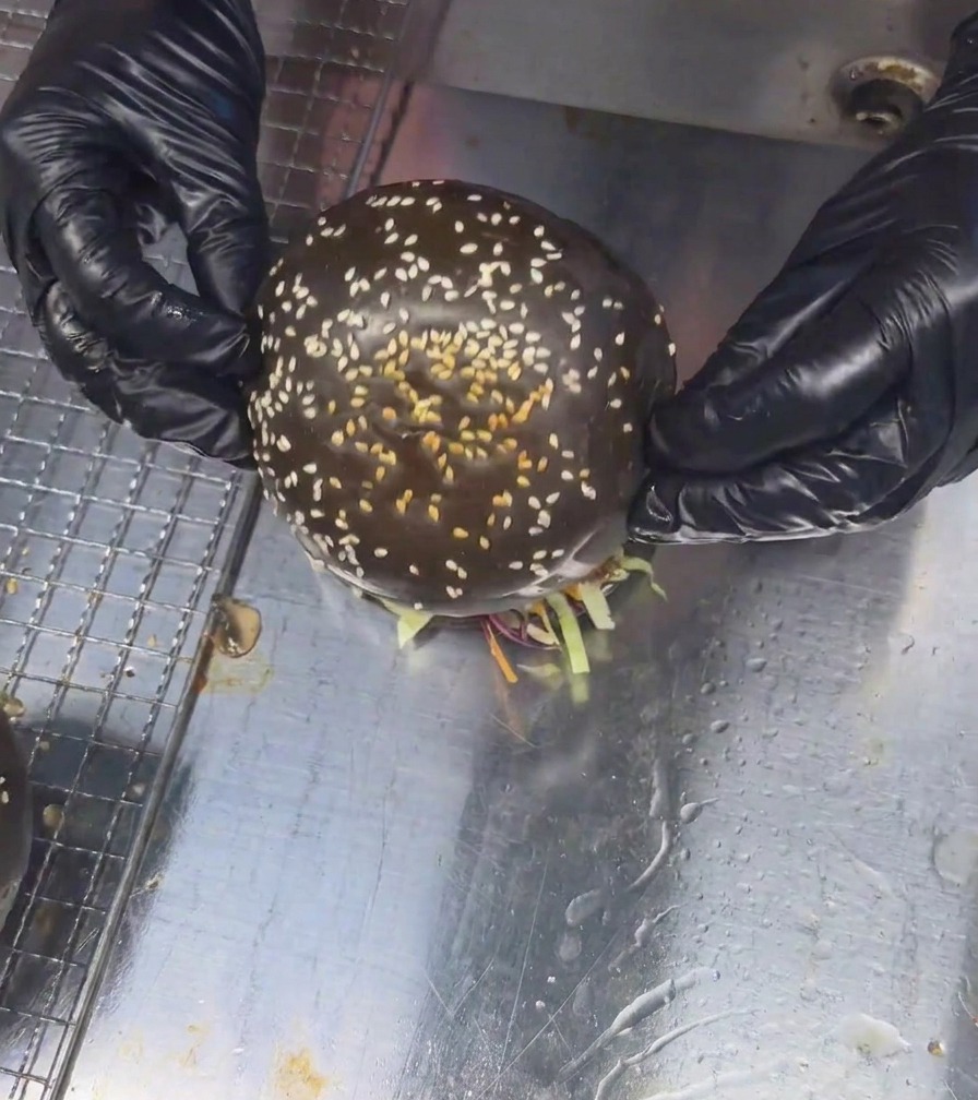 Two gloved hands placing a black sesame seed bun onto a tall fully assembled mushroom beef burger.
