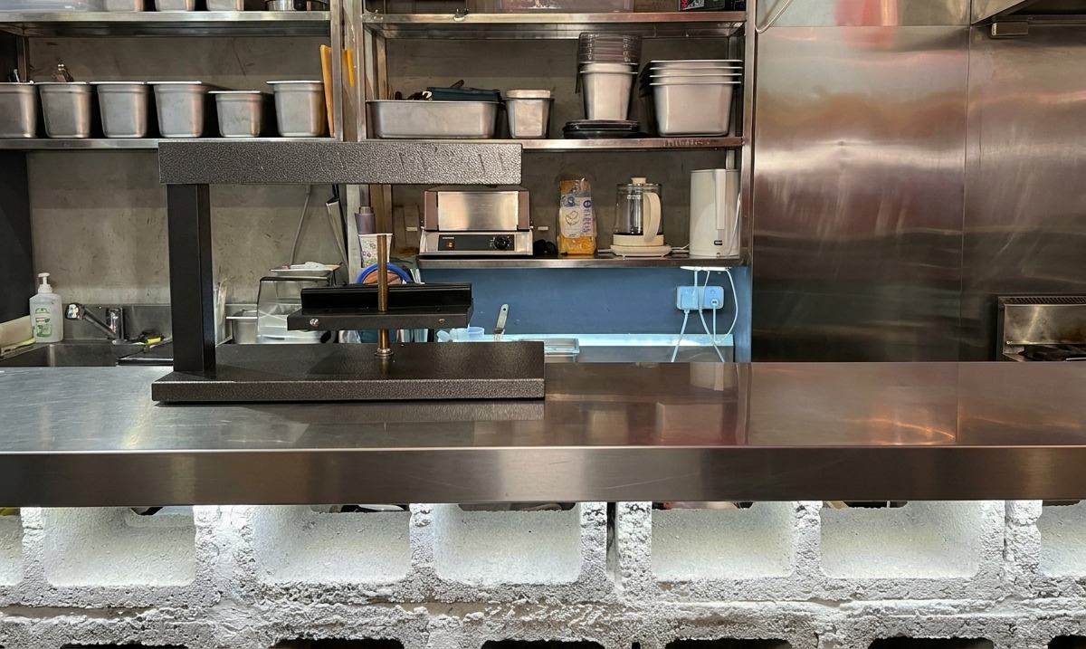 A black electric cheese raclette heater standing ready on a stainless steel commercial kitchen counter.