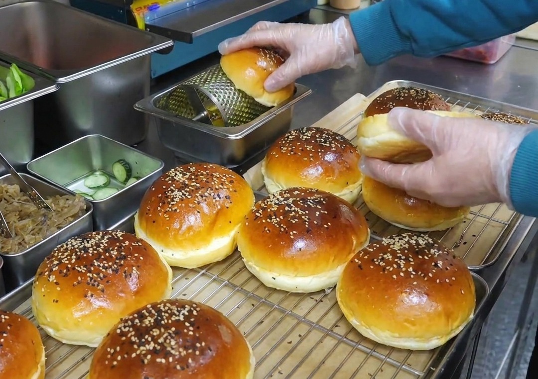 Hands in plastic gloves rolling sesame seed buns over a rotating metal butter application tool.