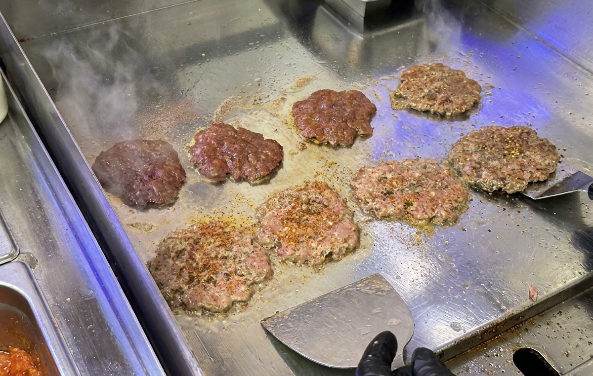 Beef burger patties being smashed and seared on a stainless steel griddle with a metal kitchen scraper.