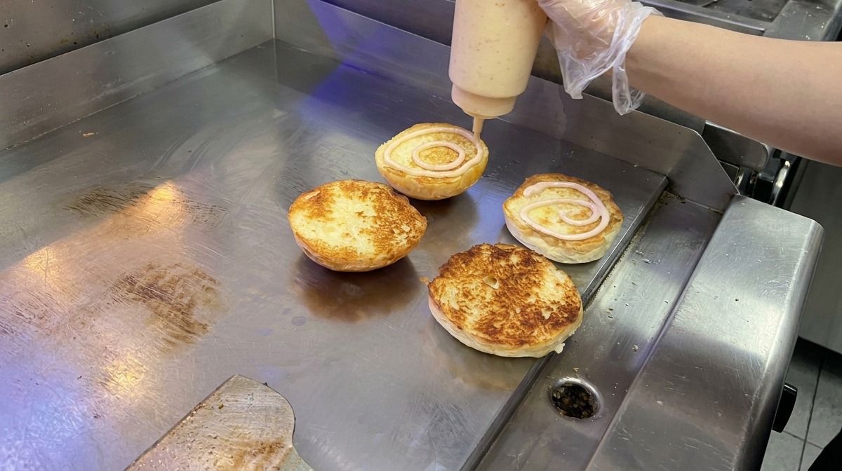 Creamy pink sauce being applied in a circular motion to toasted brioche buns on a flat-top grill.
