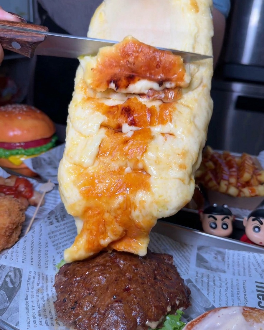 A generous layer of melted charred cheese being scraped from a large wheel with a knife and layered onto a beef burger patty
