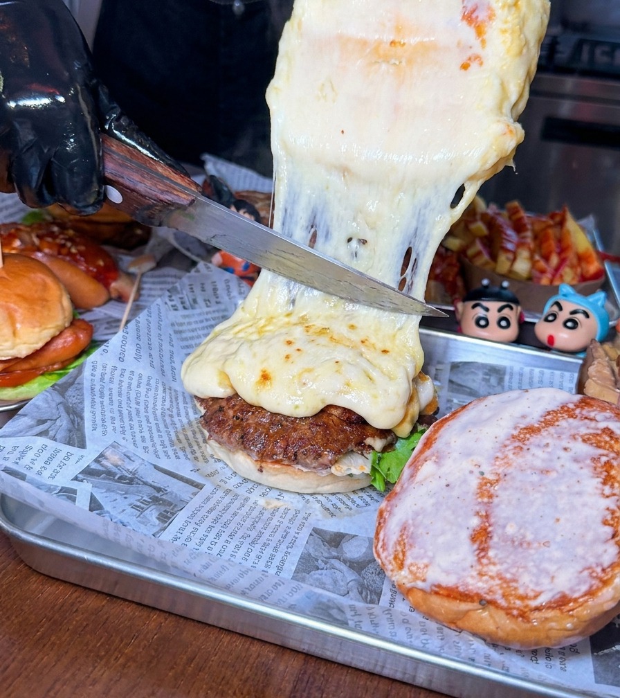 A large knife scraping a thick layer of melted, bubbly cheese from a wheel onto a beef burger patty.