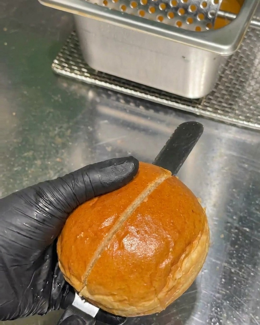 A hand in a black glove using a long serrated knife to slice a golden-brown burger bun in half on a stainless steel countertop.