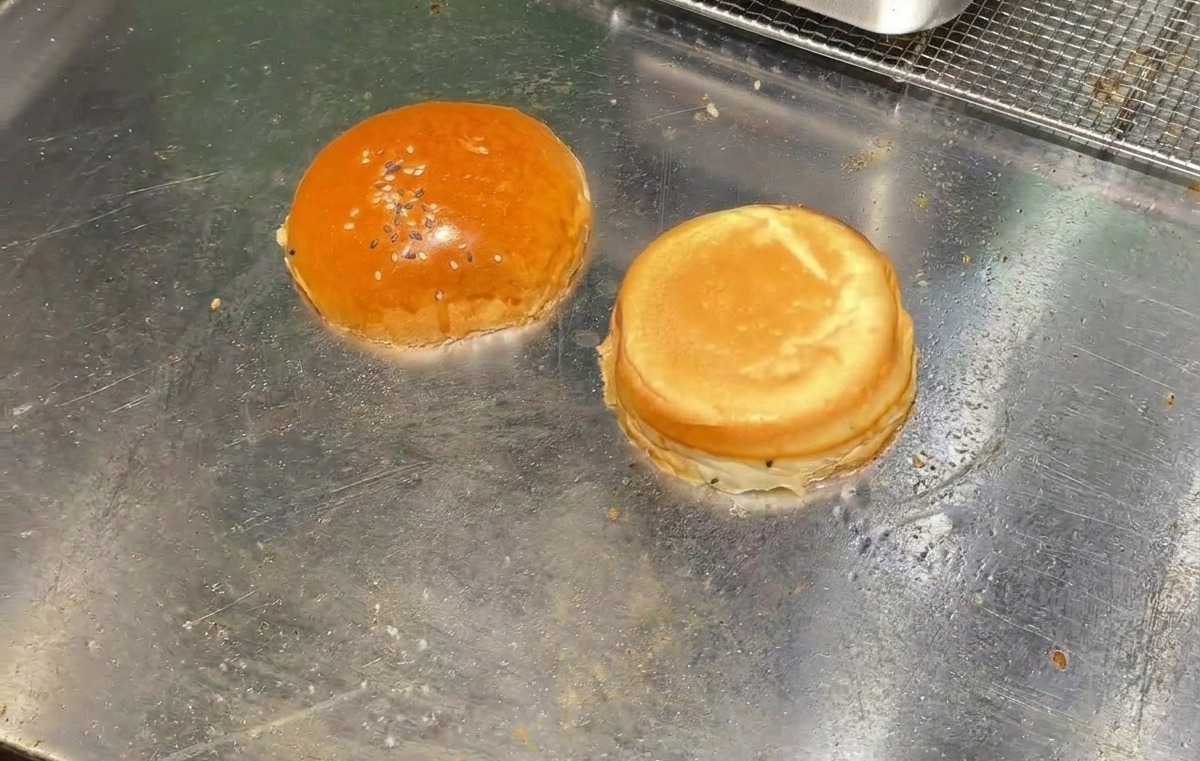 Two halves of a burger bun toasting face-down on a flat metal grill surface.