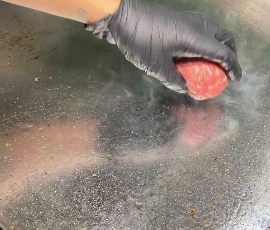 A gloved hand placing a raw, pink beef meatball onto a hot metal griddle with visible steam rising from the contact point.