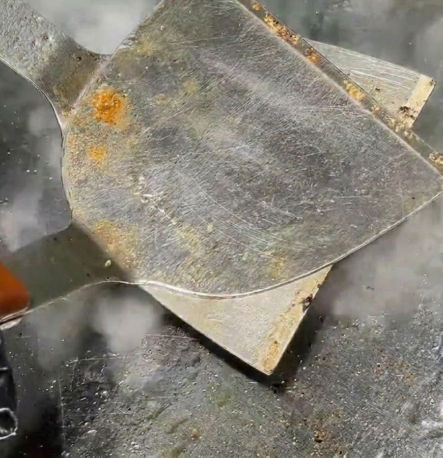 A metal spatula pressing a round meatball into a flat patty on a hot grill with steam rising.